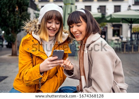 Stock photo of two caucasian girls looking at a smartphone. They are laughing. They are seated on the street. One of them is pointing at the phone.