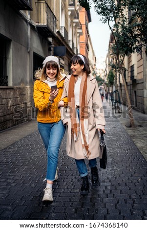 Stock photo of two caucasian girls walking in the street together laughing and having fun. They are looking at a smartphone.