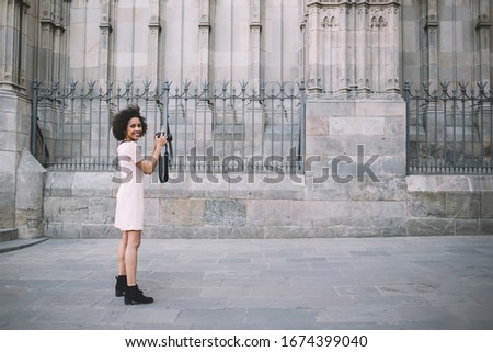 Side view of optimistic African American photographer smiling and taking pictures outside ancient building while visiting Barcelona City on summer day