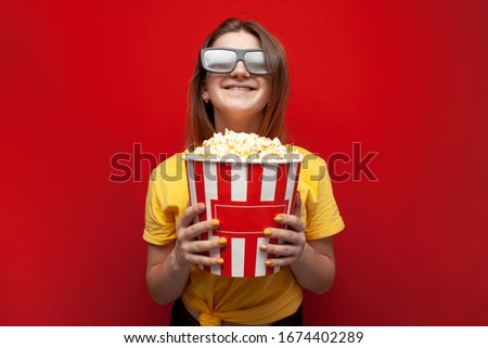 happy young girl the viewer in 3D glasses holds a big box of popcorn and watches a movie on a red color background, copy space
