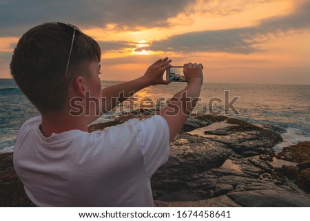 Mobile photographer. Young boy, teenager, making photo of beautiful ocean sunset on mobile phone, standing on rocky coast.
