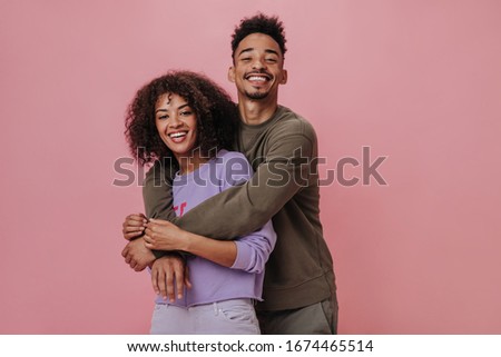 Portrait of happy couple hugging and smiling on pink background. Dark-skinned woman in purple sweater and her boyfriend posing on isolated
