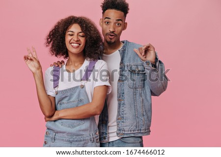 Cheerful girl showing peace sign while her boyfriend smiling on pink background. Pretty woman in white tee and man in denim jacket laugh on iolated