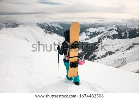 Staying snowboarder with ski poles walking along trail on the background of snow-capped mountains and cloudy sky.