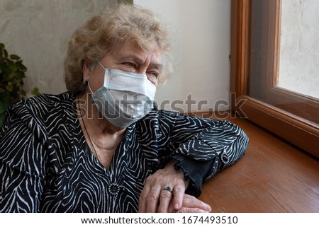 An elderly woman in a protective mask sits at home and looks out the window. Quarantine during a coronavirus pandemic. Royalty-Free Stock Photo #1674493510