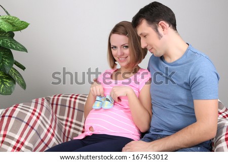 Young pregnant woman holding baby shoes with her husband on sofa at home