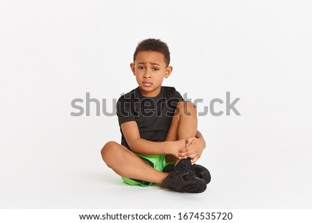 People, sports and injury concept. Picture of sad upset African little boy sitting on floor in sportswear and running shoes embracing legs, having painful facial expression because of twisted ankle