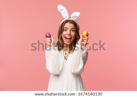 Holidays, spring and party concept. Portrait of excited charming young woman celebrating Easter in rabbit ears and white party dress, dancing with two painted eggs, pink background
