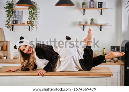 Lady dressed up in plush pajama in form of cartoon character panda is having fun at bachelorette party, posing on kitchen table, smiling. Close-up.