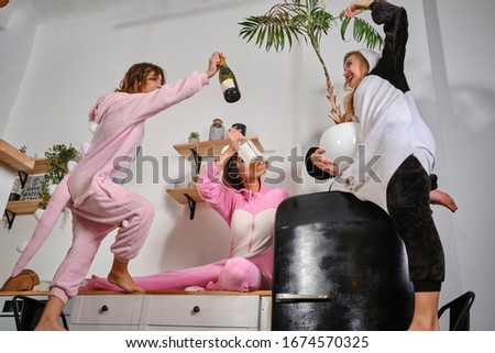 Cute maidens dressed in plush pajamas in form of cartoons characters are enjoying bachelorette party, drinking alcohol, posing on kitchen worktops.