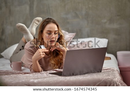 Blonde girl blogger in glasses, casual clothing. Propping her chin, holding plastic card, lying on bed with laptop, book and cup. Close-up, copy space