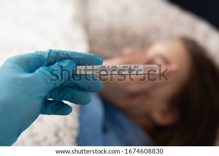 Medical thermometer being held by hand with blue rubber glove against ill blonde woman lying in bed. Coronavirus disease outbreak.