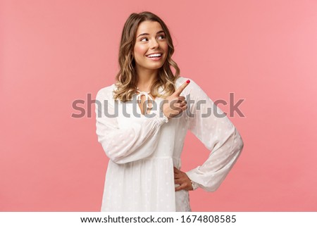 Look at this. Cheerful lovely blond european woman 20s in white cute dress, pointing at upper right corner with beaming smile, looking pleased, satisfied with good offer, pink background