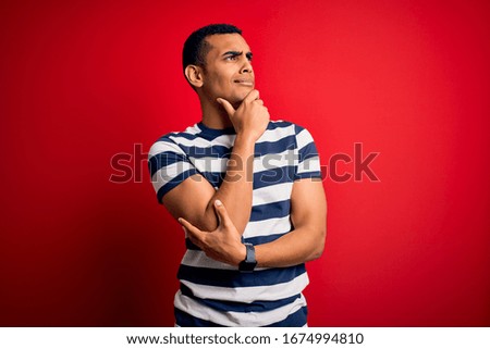 Handsome african american man wearing casual striped t-shirt standing over red background Thinking worried about a question, concerned and nervous with hand on chin
