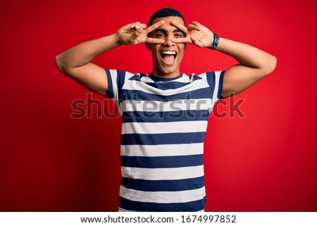 Handsome african american man wearing casual striped t-shirt standing over red background Doing peace symbol with fingers over face, smiling cheerful showing victory