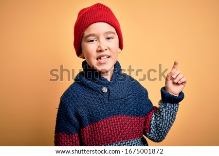 Young little boy kid wearing wool cap and winter sweater over yellow isolated background with a big smile on face, pointing with hand and finger to the side looking at the camera.