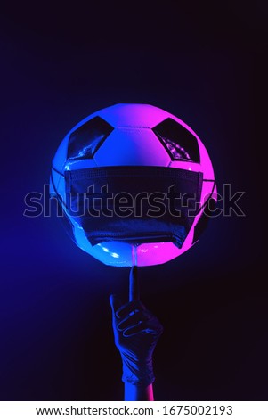 A soccer ball in a black medical mask from the virus. Hand in glove with finger raised. In the light of neon on a dark background.