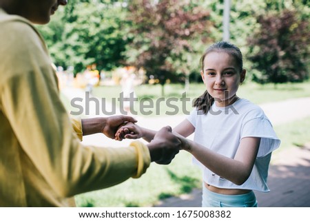 Girl in casual clothes holding hands of brother and looking at camera while learning to ride skates on blurred background of park