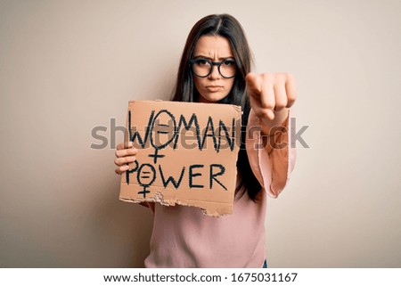 Young brunette woman holding women power protest banner over isolated background pointing with finger to the camera and to you, hand sign, positive and confident gesture from the front