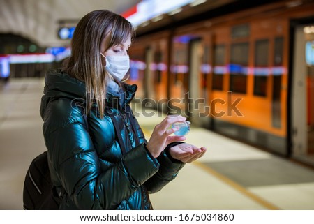 Woman in winter coat with protective mask on face standing on metro station, using hand sanitizer, looking worried. Preventive measures in public places of epidemic regions. Finland, Espoo Royalty-Free Stock Photo #1675034860