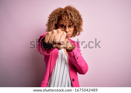 Young african american businesswoman wearing glasses standing over pink background Punching fist to fight, aggressive and angry attack, threat and violence