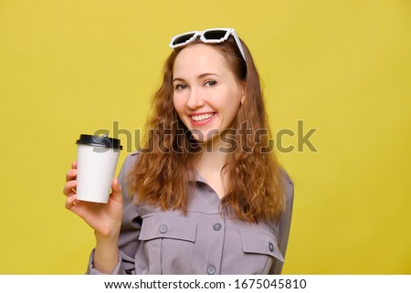 Stylish caucasian girl in a gray dress on a yellow background. Holding a disposable coffee cup to go. Copy space. Close up.