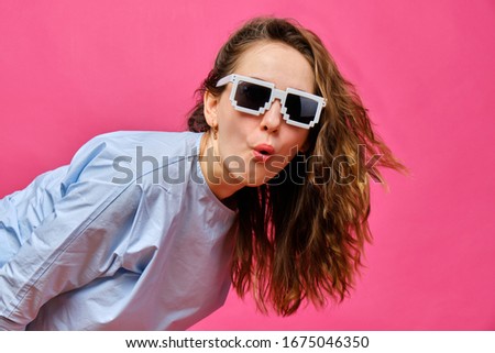 Stylish caucasian girl in a pale blue t-shirt and 8-bit glasses and makes a surprised look on a pink background. Close up.
