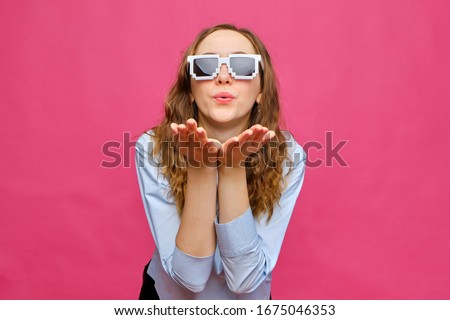 Stylish caucasian girl in a pale blue t-shirt and 8-bit glasses and blows a kiss on a pink background. Close up.