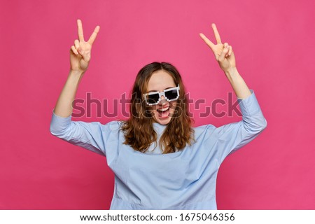 Stylish caucasian girl in a pale blue t-shirt and 8-bit glasses shows fingers abstract peace hand and shouts smiling on a pink background. Close up.