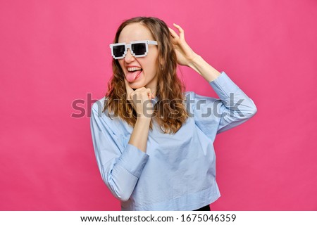 Stylish caucasian girl in a pale blue t-shirt and 8-bit glasses shows tongue and grimaces on a pink background. Close up.