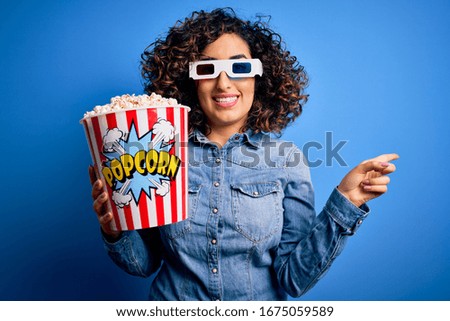 Young beautiful curly arab woman watching movie using 3d glasses eating popcorn snack very happy pointing with hand and finger to the side