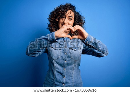 Young beautiful curly arab woman wearing casual denim shirt standing over blue background smiling in love doing heart symbol shape with hands. Romantic concept.