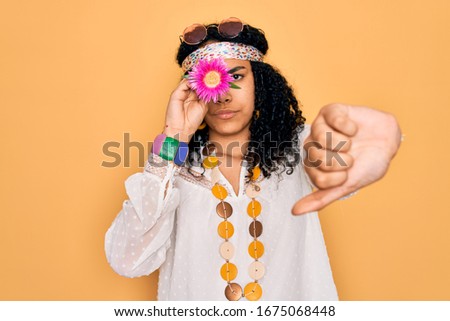Young african american curly hippie woman wearing sunglasses holding pink flower on eye with angry face, negative sign showing dislike with thumbs down, rejection concept