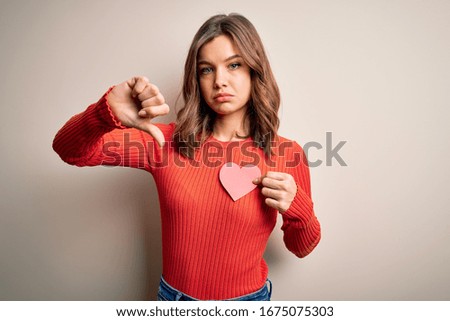 Young blonde romantic girl holding red heart paper shape over isolated background with angry face, negative sign showing dislike with thumbs down, rejection concept
