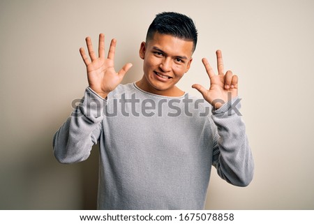Young handsome latin man wearing casual sweater standing over isolated white background showing and pointing up with fingers number eight while smiling confident and happy.