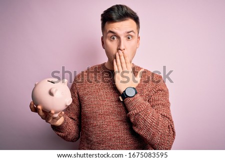 Young handsome caucasian man holding piggy bank for savings over pink background cover mouth with hand shocked with shame for mistake, expression of fear, scared in silence, secret concept Royalty-Free Stock Photo #1675083595