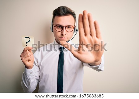 Young call center operator man wearing headset holding paper note with question mark with open hand doing stop sign with serious and confident expression, defense gesture