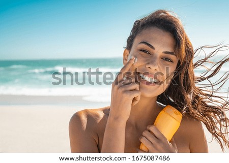 Beautiful young woman at beach applying sunscreen on face and looking at camera. Beauty latin girl applying suntan lotion at sea. Portrait of happy woman with healthy skin applying sunblock on cheek. Royalty-Free Stock Photo #1675087486
