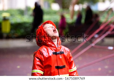 Funny child dressed in red raincoat firefighter play in a park on a rainy day.