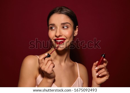 Positive young charming brown-eyed dark haired woman with evening makeup holding red lip gloss in raised hand and smiling cheerfully while posing over burgundy background