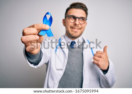 Young doctor man wearing medical coat and holding colon cancer awareness blue ribbon pointing and showing with thumb up to the side with happy face smiling