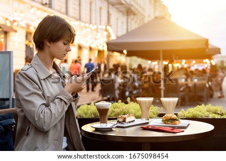Millennial hipster teen girl blogger hold smart phone take food photo on phone mobile camera sit in city cafe at table. Young female vlogger shoot social media video story blog on smartphone outdoor