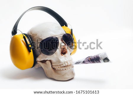 A human skull wearing yellow headphones and dark glasses holds a 500 Euro bill rolled into a cigarette between its teeth. The concept of the danger and cost of tobacco Smoking and drug addiction.