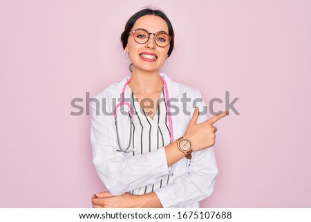 Beautiful doctor woman with blue eyes wearing coat and stethoscope over pink background cheerful with a smile of face pointing with hand and finger up to the side with happy and natural expression 