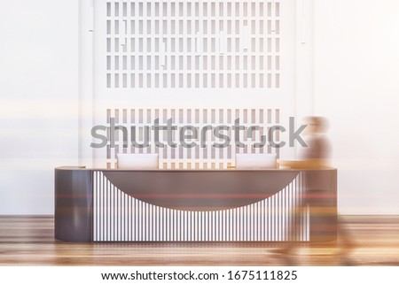Blurry businessman passing by gray and white reception desk in modern office hall with white walls and wooden floor. Toned image