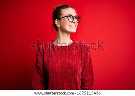 Young beautiful redhead woman wearing casual sweater over isolated red background looking away to side with smile on face, natural expression. Laughing confident.