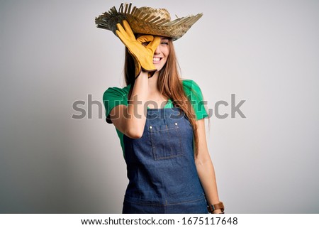 Young beautiful redhead farmer woman wearing apron and hat over white background doing ok gesture with hand smiling, eye looking through fingers with happy face.