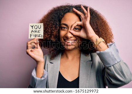 Young african american woman with afro hair holding paper with you are fired message with happy face smiling doing ok sign with hand on eye looking through fingers