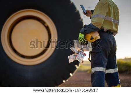 Haul truck inspector wearing work uniform safety glove holding hard hat danger tags personal locks inspection pre operation book inspecting taking pic with cell phone defocused haul truck background Royalty-Free Stock Photo #1675140619