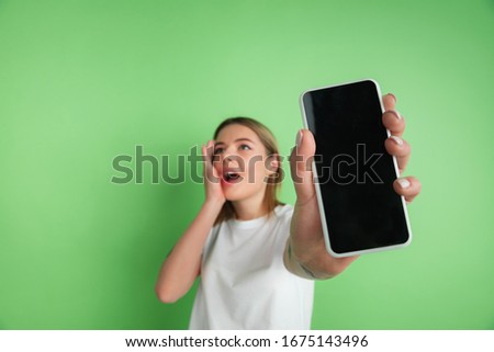 Showing blank screen. Caucasian young woman's portrait isolated on green studio background. Beautiful female model in white shirt. Concept of human emotions, facial expression, sales, ad, youth.